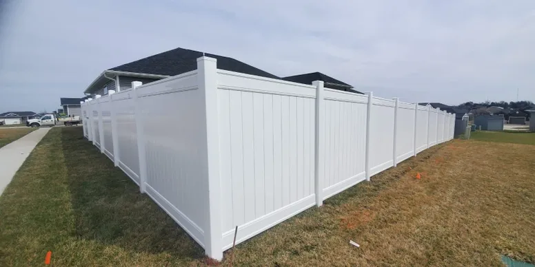 A white vinyl privacy fence surrounds the backyard of a suburban house, with grass and a sidewalk visible in the foreground.