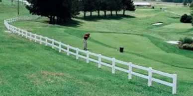 A person stands on a golf course preparing to take a shot near a white fence, with trees and grassy areas in the background.