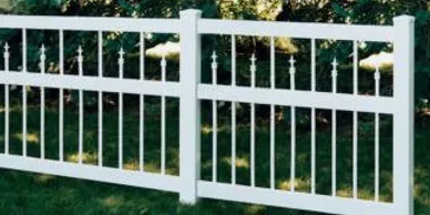 White metal fence panels with vertical bars and decorative finials stand on grass with bushes in the background.