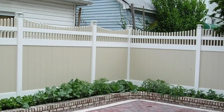 A beige and white vinyl fence encloses a backyard garden with green plants bordered by a low brick wall.