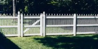 A white picket fence with a gate stands on a lawn, with trees and bushes visible in the background.