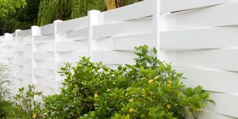 A white vinyl privacy fence with horizontal panels stands behind green shrubs and foliage.