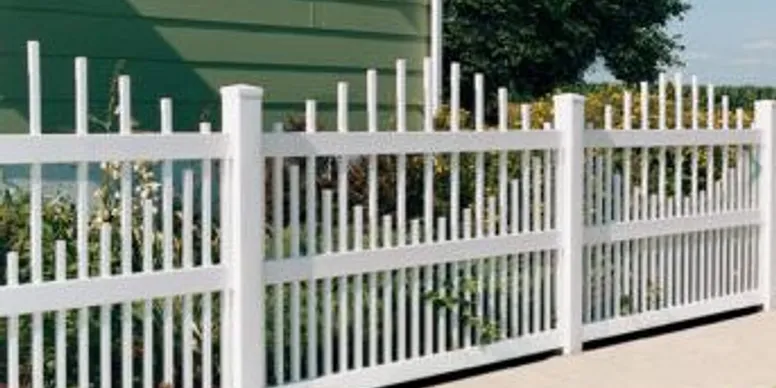 A white picket fence with vertical slats of varying heights stands in front of a green building and some plants.