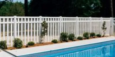 A white picket fence runs alongside a pool, with small shrubs and plants growing at the base of the fence.