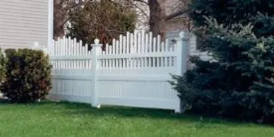 A white vinyl picket fence with decorative posts stands on a green lawn, bordered by a bush on the left and an evergreen tree on the right.