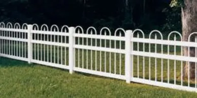 A white metal fence with arched tops stands on green grass beside a tree, with a dark background of foliage.