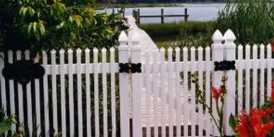 A white picket fence with a gate is in the foreground, with greenery, red flowers, and water with a dock visible in the background.