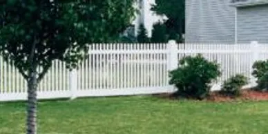 A white picket fence surrounds a grassy yard with a tree and several bushes near a house.