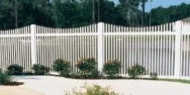 White vinyl picket fence with a scalloped design encloses a yard; small bushes are planted along the inside of the fence, and trees are visible in the background.