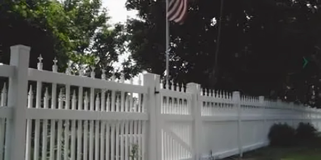 A white picket fence runs alongside a yard with trees and an American flag on a pole in the background.