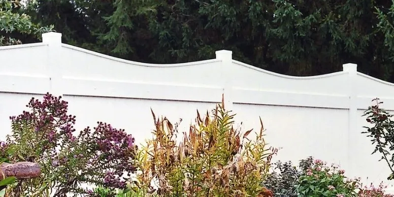 A white vinyl privacy fence with a scalloped top stands behind a garden with various green plants and purple flowers.
