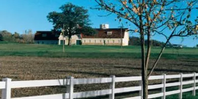 A white fence runs along a harvested field; a tree stands nearby, and a large farmhouse with a red roof sits in the background under a clear blue sky.