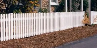 A white picket fence runs alongside a mulched area and a paved road, with a house and some vegetation in the background.