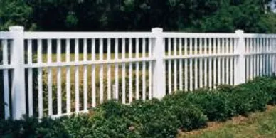 A white wooden picket fence runs along a grassy yard with green shrubs planted in front of it.