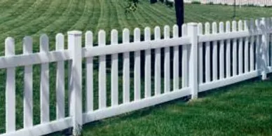 A white picket fence runs alongside a green, neatly mowed lawn with trees in the background.