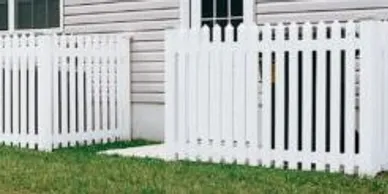 A white picket fence runs along the side of a house with beige siding and a window, standing on a small patch of grass.