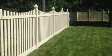 A cream-colored vinyl picket fence with pointed tops runs alongside a neatly mowed green lawn in a yard.
