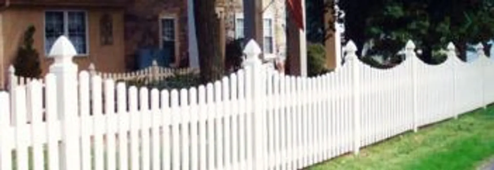 A white picket fence with pointed tops runs along the edge of a green lawn in front of a house.