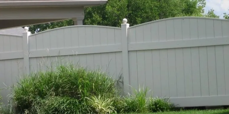 A white vinyl privacy fence with decorative posts stands behind a patch of tall green grass and shrubs.