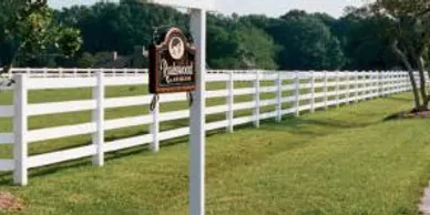 A long white wooden fence runs alongside a grassy lawn with a hanging wooden sign attached to a white post in the foreground. Trees are visible in the background.