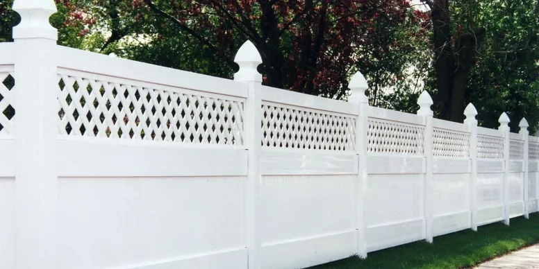 A long white vinyl privacy fence with decorative lattice panels on top, set next to trees and grass.