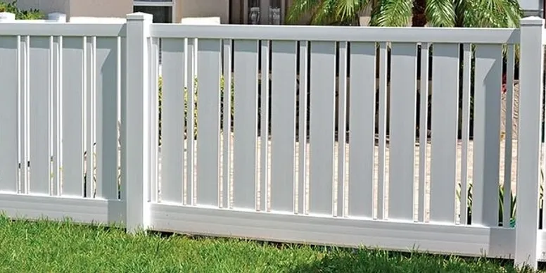 A white vinyl fence with vertical slats stands on green grass, with a house and palm trees visible in the background.