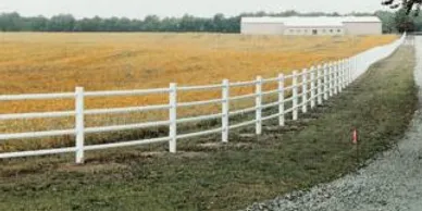 A white wooden fence runs alongside a gravel road next to a field with a large building in the background.