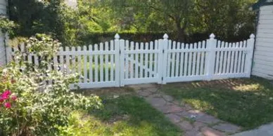 A white picket fence with a gated entrance stands between a grassy yard and a stone pathway, bordered by plants and a building.