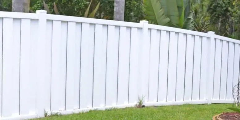 A white vinyl privacy fence with vertical panels stands on a green lawn with trees and plants in the background.