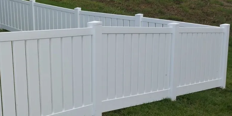 White vinyl fencing sections are installed on a grassy slope, forming angled boundaries with vertical panel designs and capped posts.