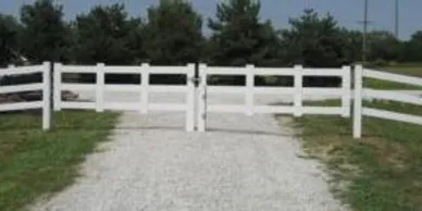 A closed white wooden gate blocks a gravel driveway, with green grass and trees in the background.