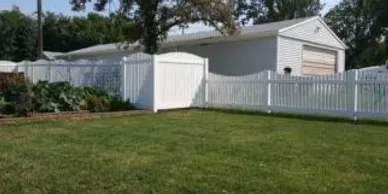 A white vinyl fence encloses a green yard with a garden on the left and a white garage in the background under a clear sky.