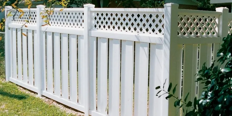 A white vinyl privacy fence with decorative lattice panels on top, installed on a grassy area with some plants nearby.