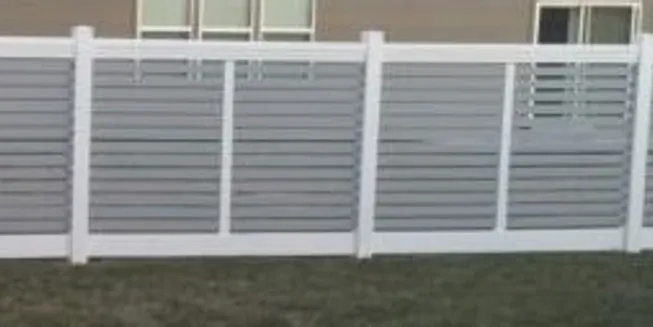 A gray and white slatted fence stands on grass in front of a building with windows and a door.