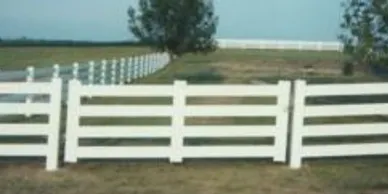 A white wooden fence runs across a grassy field, with a tree and open land visible in the background.