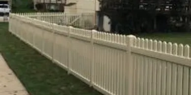 A long, white picket fence runs parallel to a sidewalk, separating a grassy yard from the walkway in a residential area.