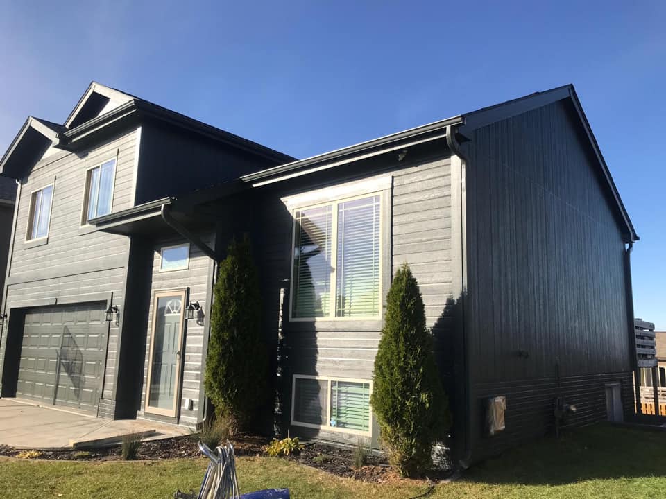 Two-story modern house with dark gray siding, large windows, and attached double garage. The lawn is mowed and there are a few small trees and shrubs in the front yard.