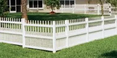 White picket fence enclosing a grassy yard with trees and a building in the background.
