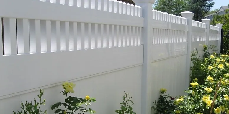 A white vinyl privacy fence with decorative lattice top panels borders a garden with yellow flowering plants.