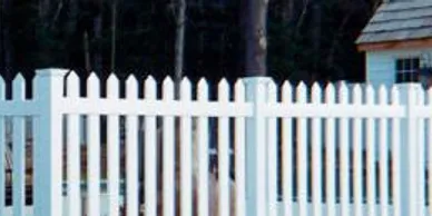 A white picket fence in front of a small building with a shingled roof, set against a backdrop of trees.