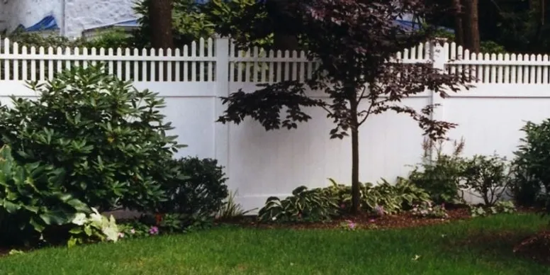 A white picket fence borders a landscaped yard with green grass, shrubs, and a small tree in front of it.