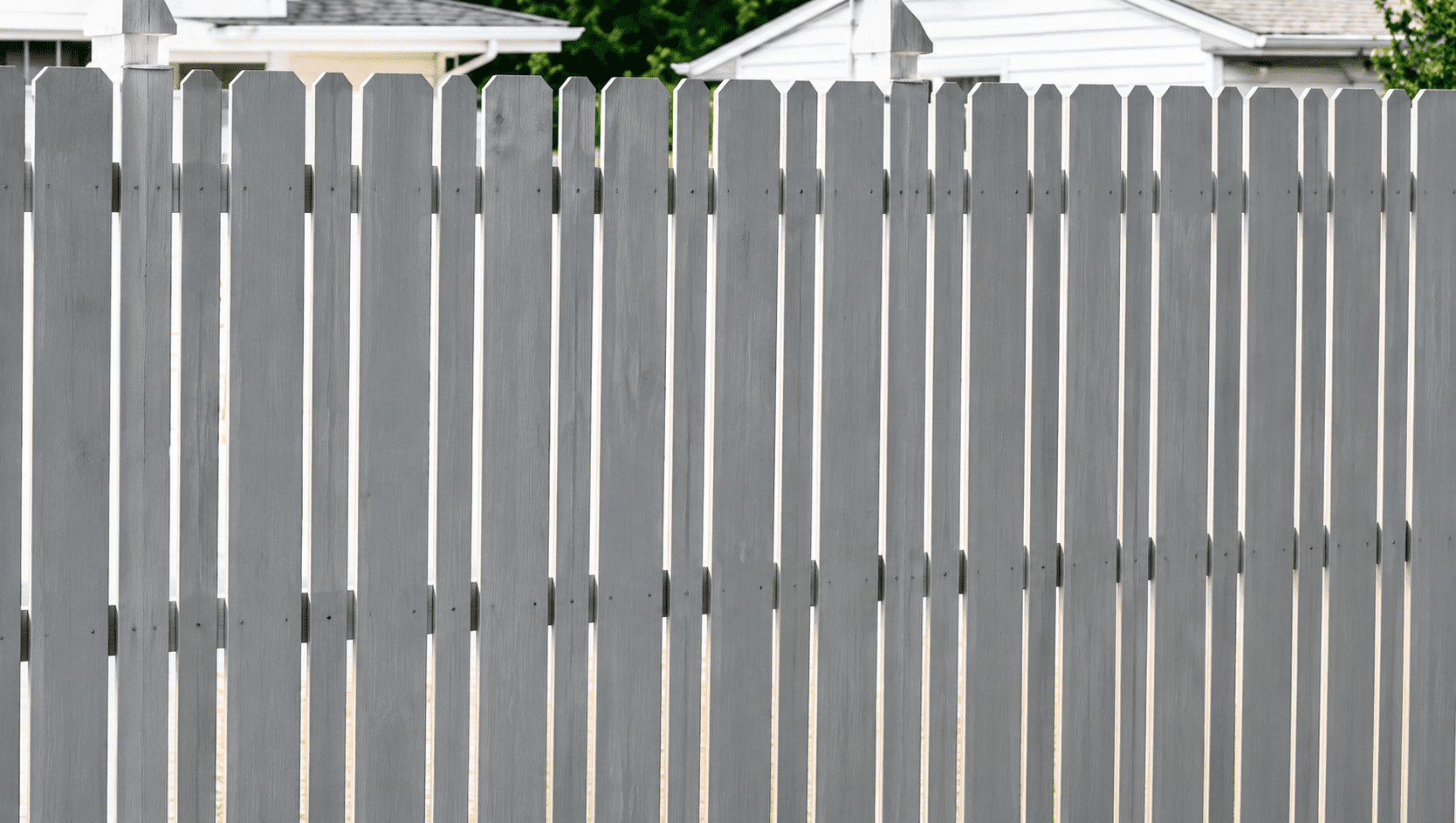 A white wooden picket fence with vertical slats stands in front of residential houses on a sunny day.
