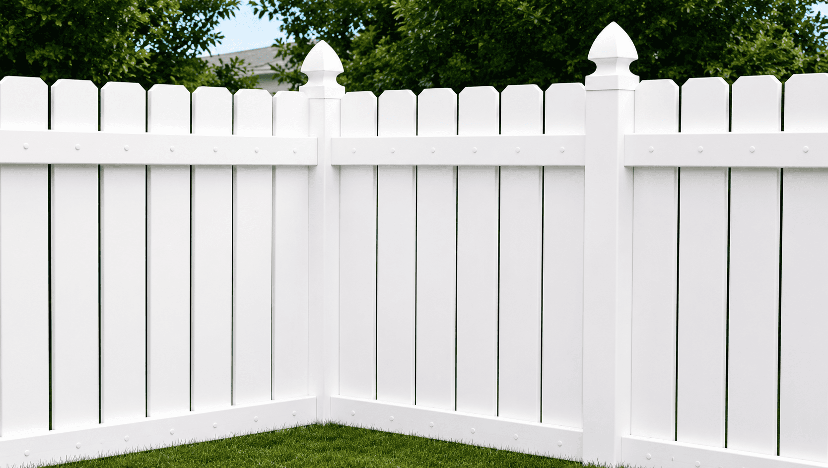 A white vinyl privacy fence forms a corner in a yard with green grass and leafy trees visible in the background.