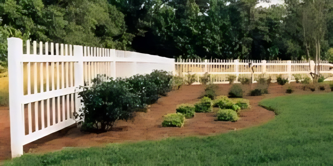 A white wooden fence runs along a garden with bushes and small shrubs, bordered by grass and trees in the background.