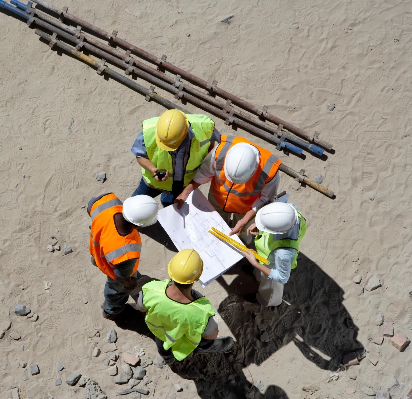 A group of five construction workers in safety gear stand on a sandy site, reviewing blueprints and documents together, demonstrating their customer-first approach to ensuring every project meets client needs.