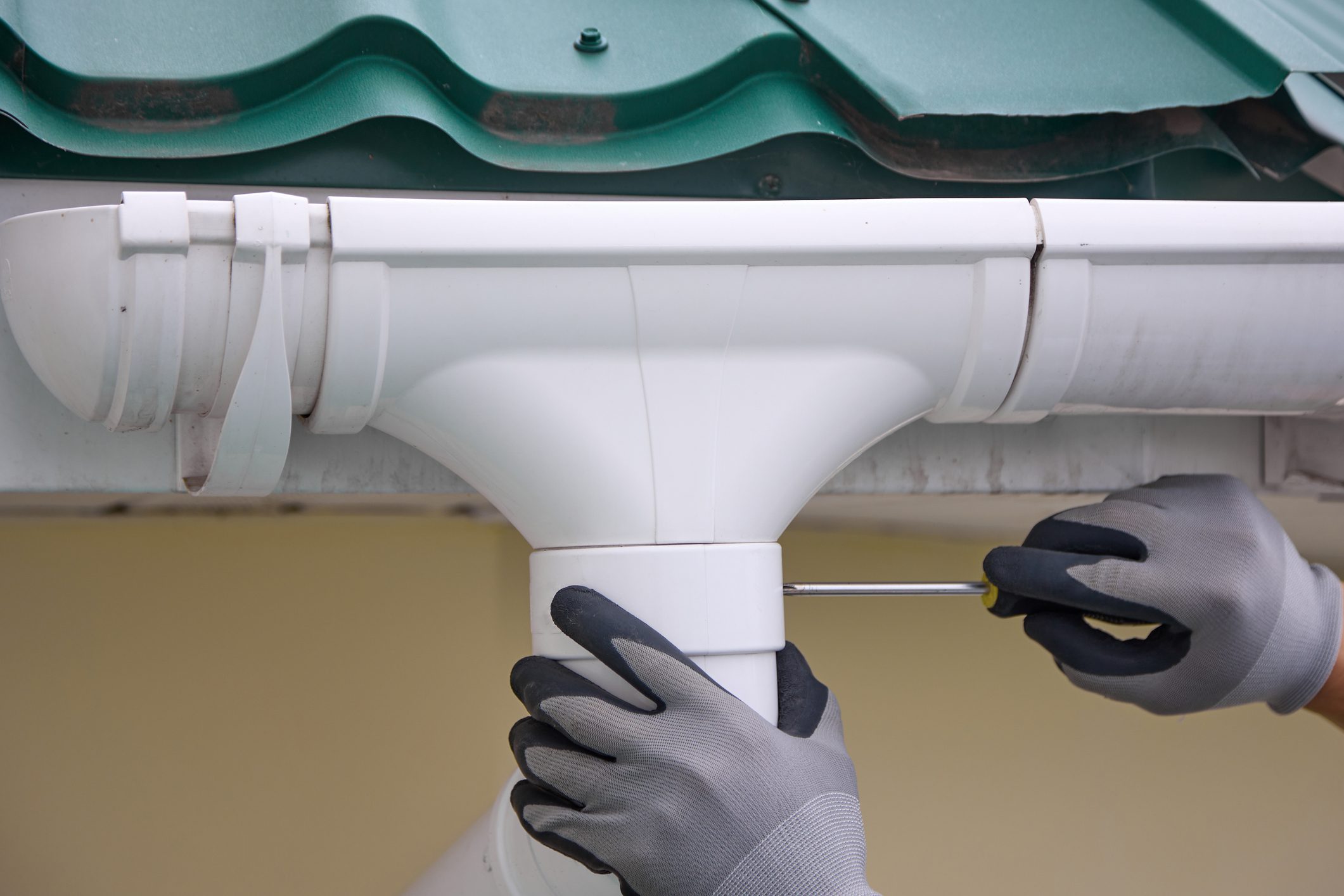 Worker in gray gloves uses a screwdriver to screw a white gutter. Builder installs a trough system on the roof to drain rainwater
