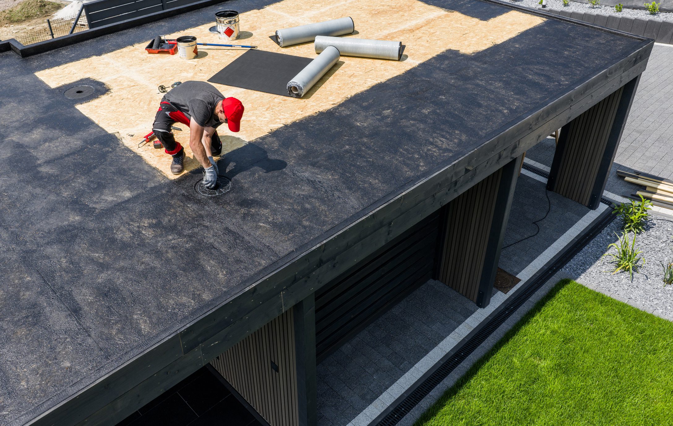 A construction worker is installing EPDM roofing material on a contemporary home under clear skies, surrounded by tools and prepared materials for the roofing project.