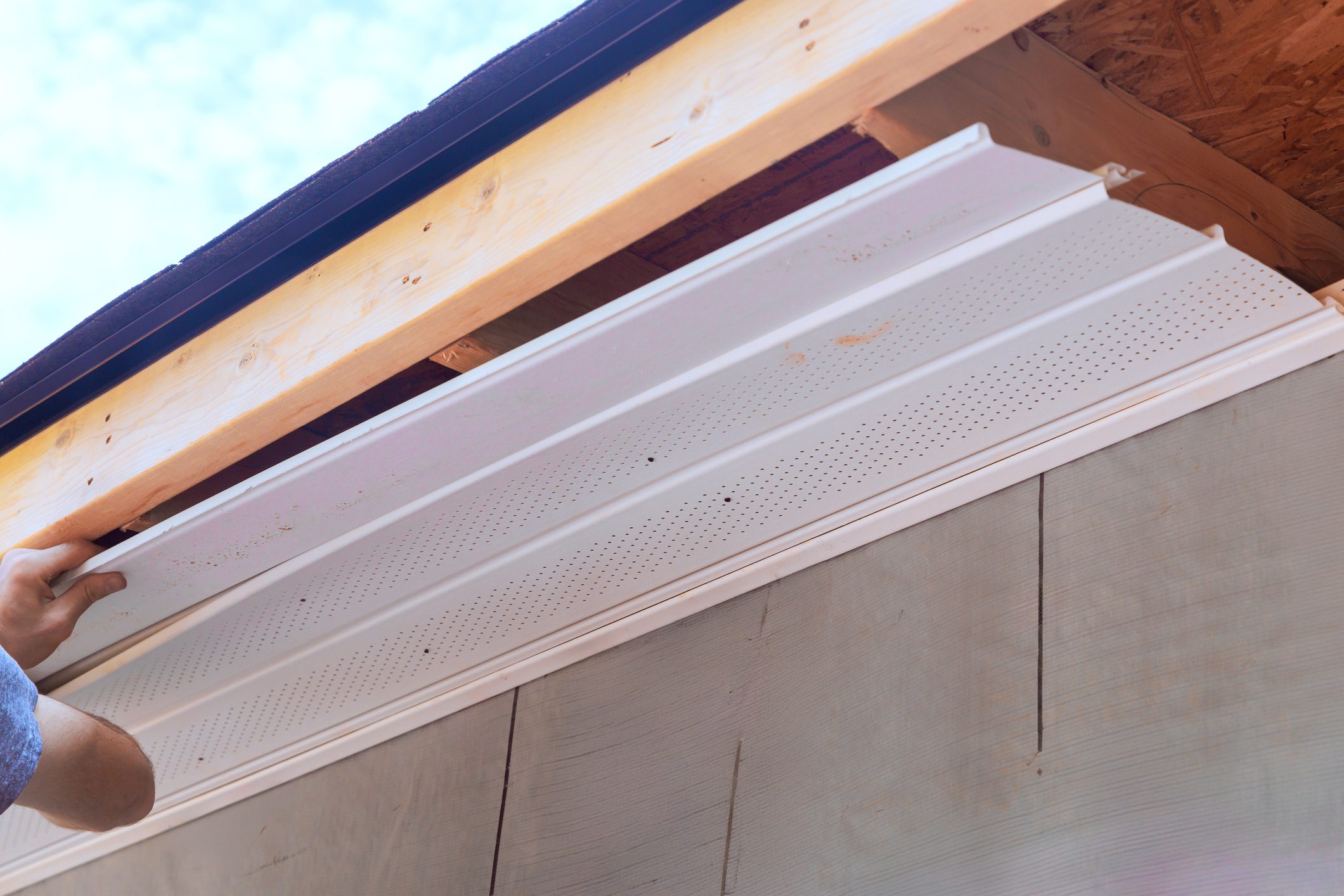 Contractor places white soffit PVC panels onto roof overhang of building while working on construction project.