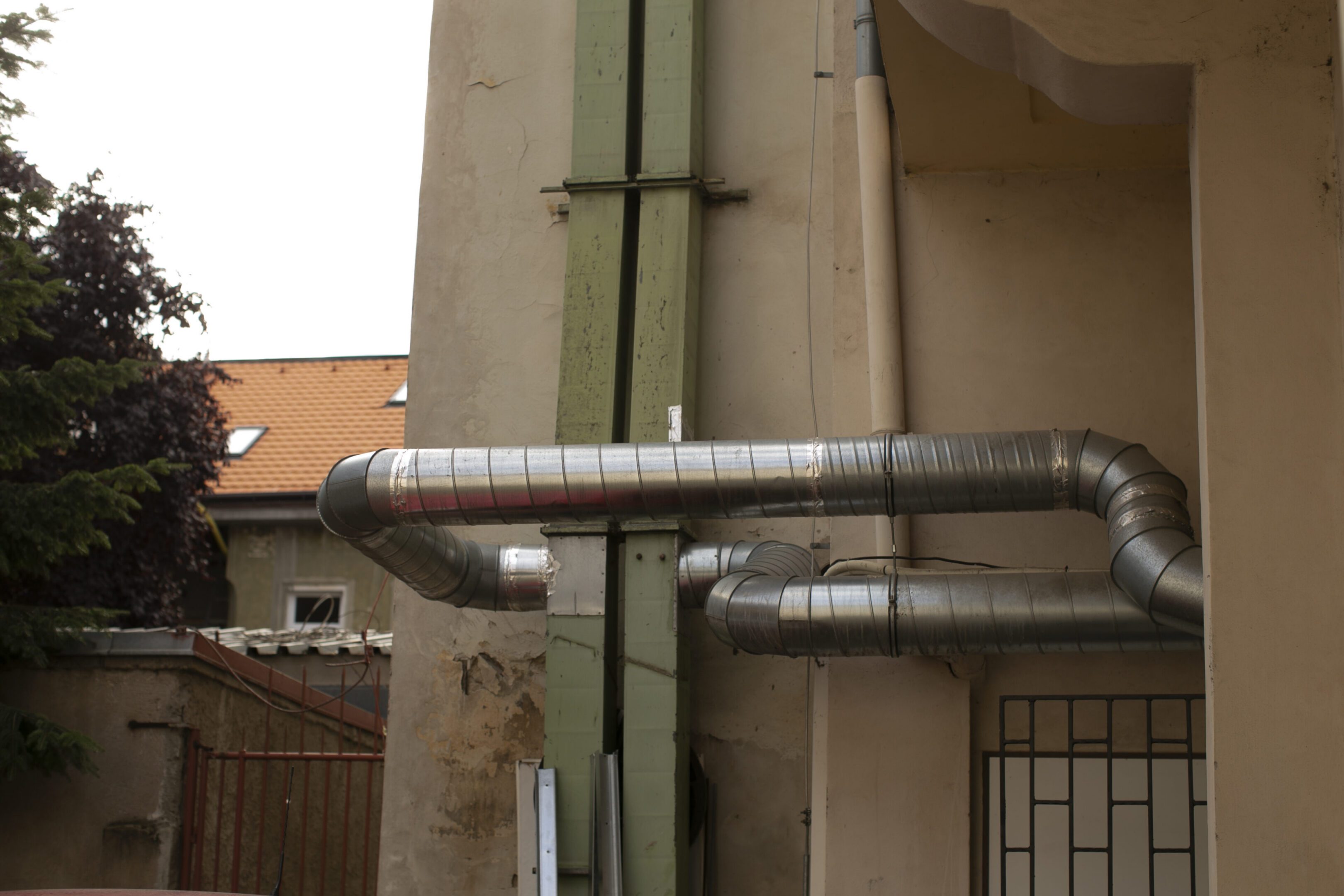 Metal ducts on building wall, urban courtyard scene showing exposed ventilation pipes and silver elbow joints attached to worn stucco facade, narrow