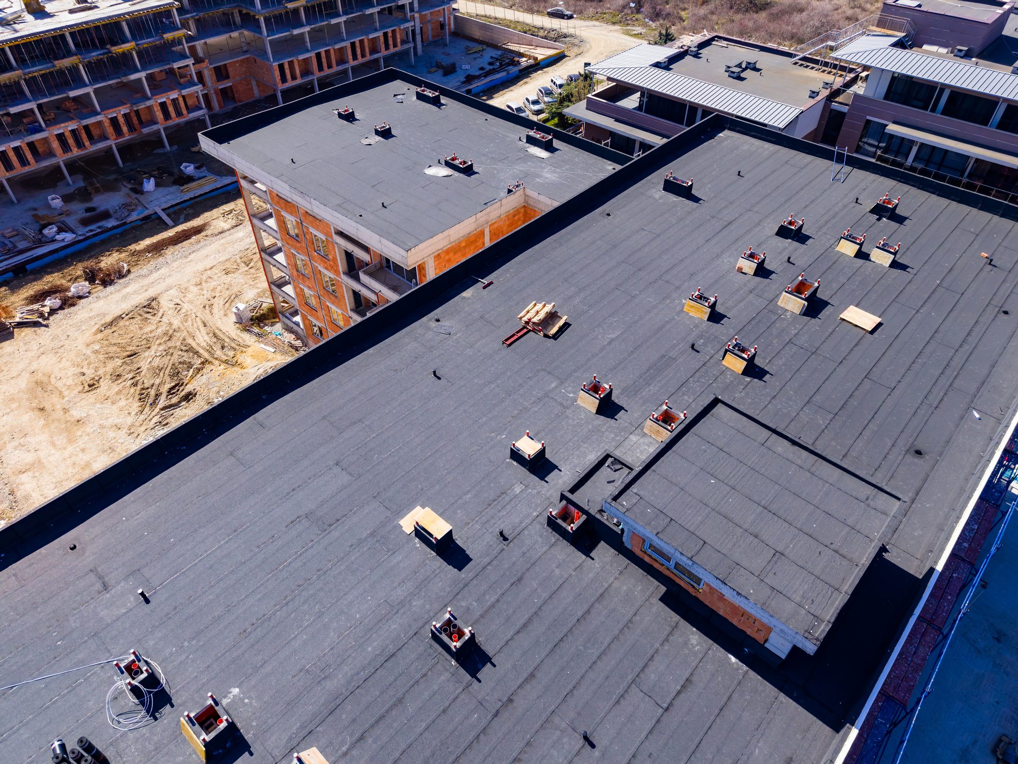 Workers are installing a new roof on a building at a construction site. There are materials placed on the rooftop. The scene shows active construction during daylight.